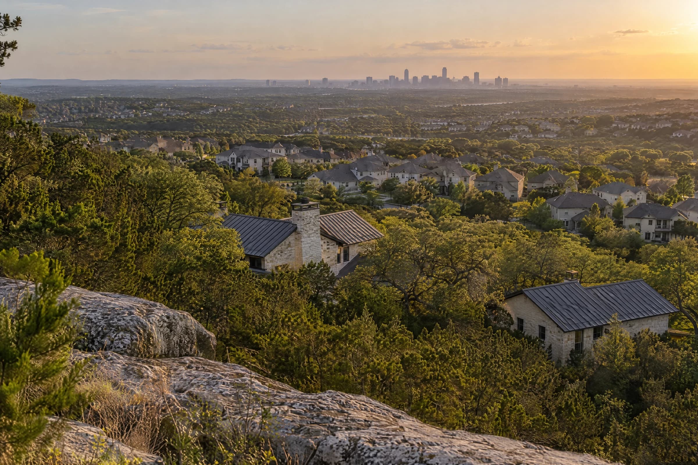 Late-afternoon view from a limestone outcrop on a cedar-covered ridge west of Austin, over Hill Country stone houses and suburban rooftops to a distant skyline