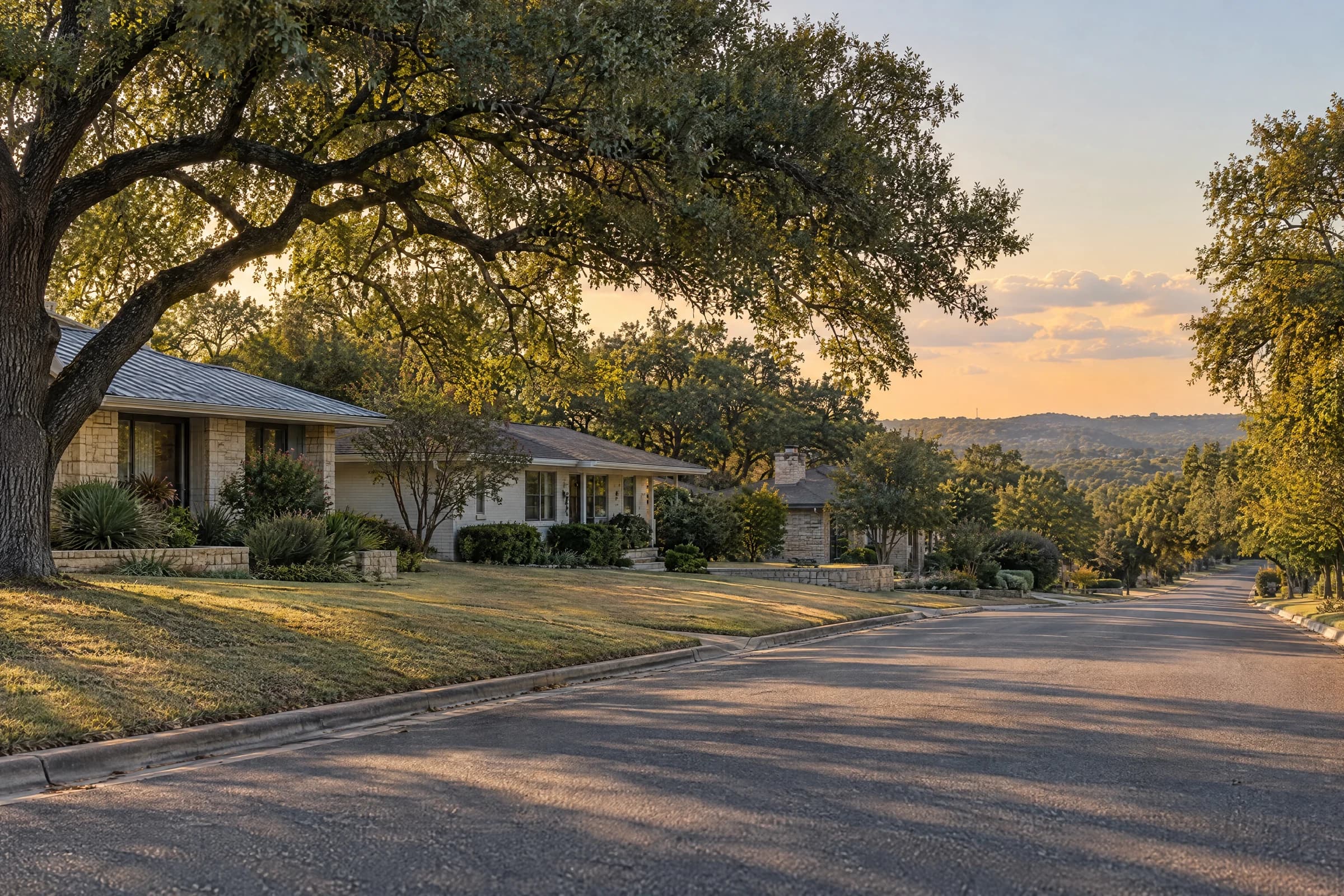 A quiet street of single-story limestone and brick ranch homes under live oaks in late-afternoon Austin light, with a Hill Country ridge in the distance