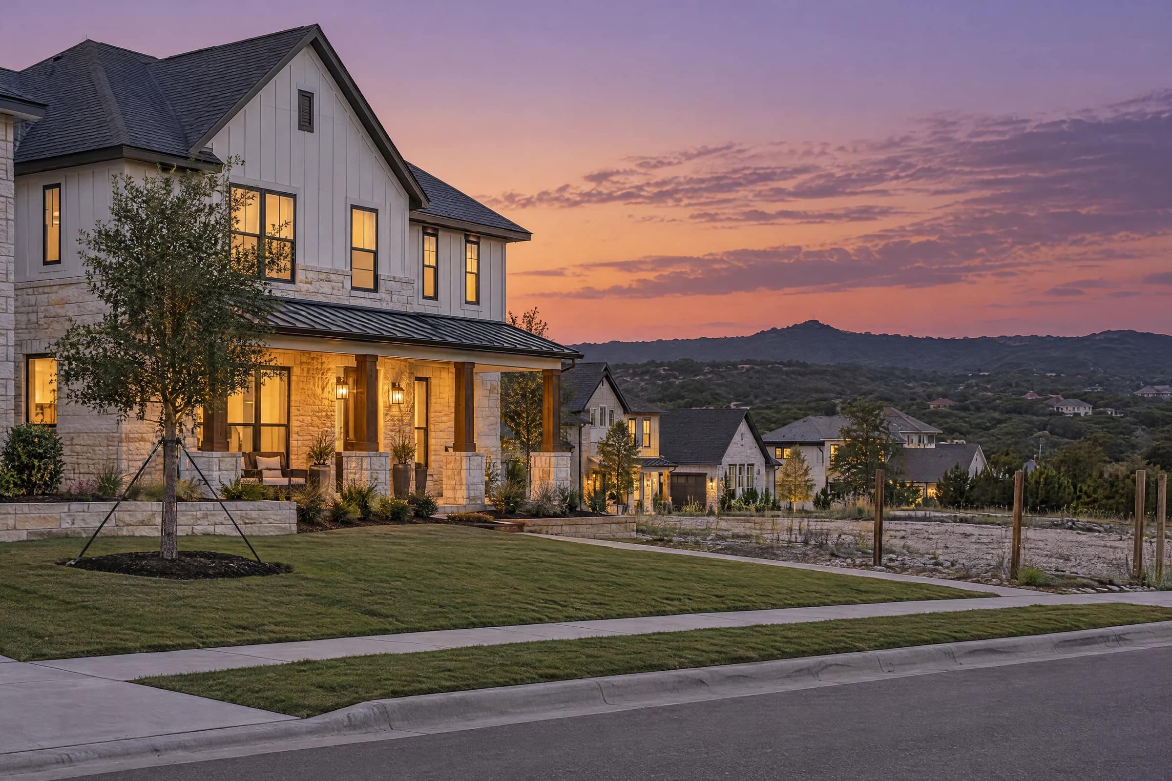 A new two-story limestone and board-and-batten home at dusk on a just-finished street north of Austin, porch lights on, a staked young live oak in the sod and empty homesites running toward a Hill Country ridge.