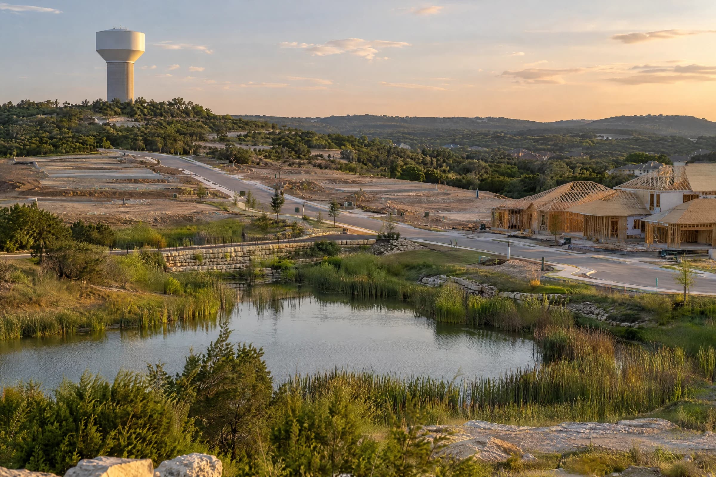 A white municipal water tower above a Hill Country subdivision under construction in Williamson County, new curbed streets on one side and framed houses on the other, cedar ridges behind