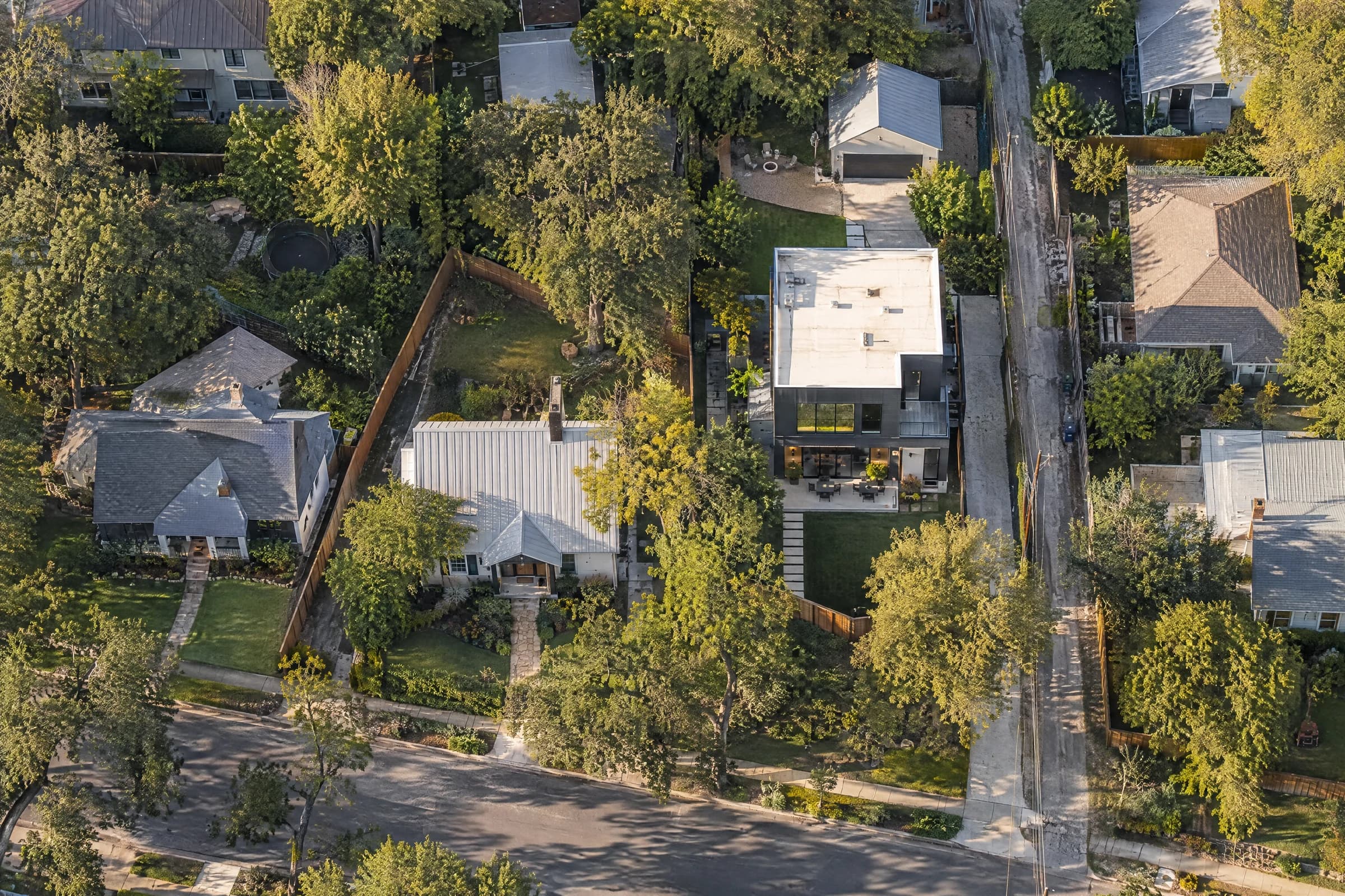 Aerial view at golden hour over a Central Austin block, fence lines tracing each lot between a metal-roofed cottage, a flat-roofed modern infill house and shingled 1950s homes under live oaks