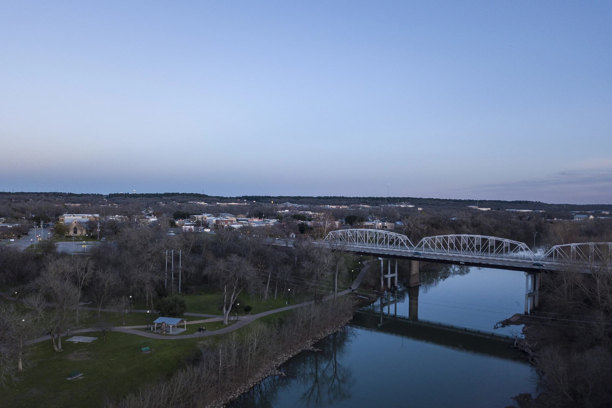 Aerial view at dusk of the truss bridges crossing the Colorado River into downtown Bastrop, Texas, with a riverside park and trail on the near bank