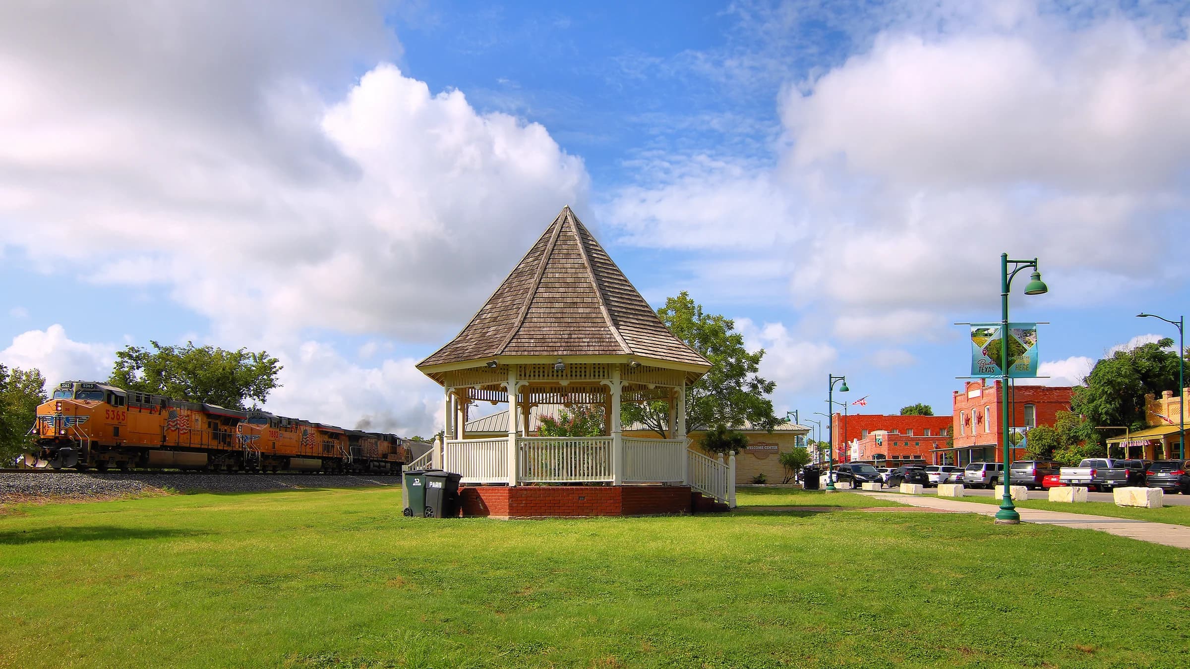 The white downtown gazebo on the green in Buda, Texas, with a Union Pacific freight train passing on one side and Main Street's brick storefronts on the other