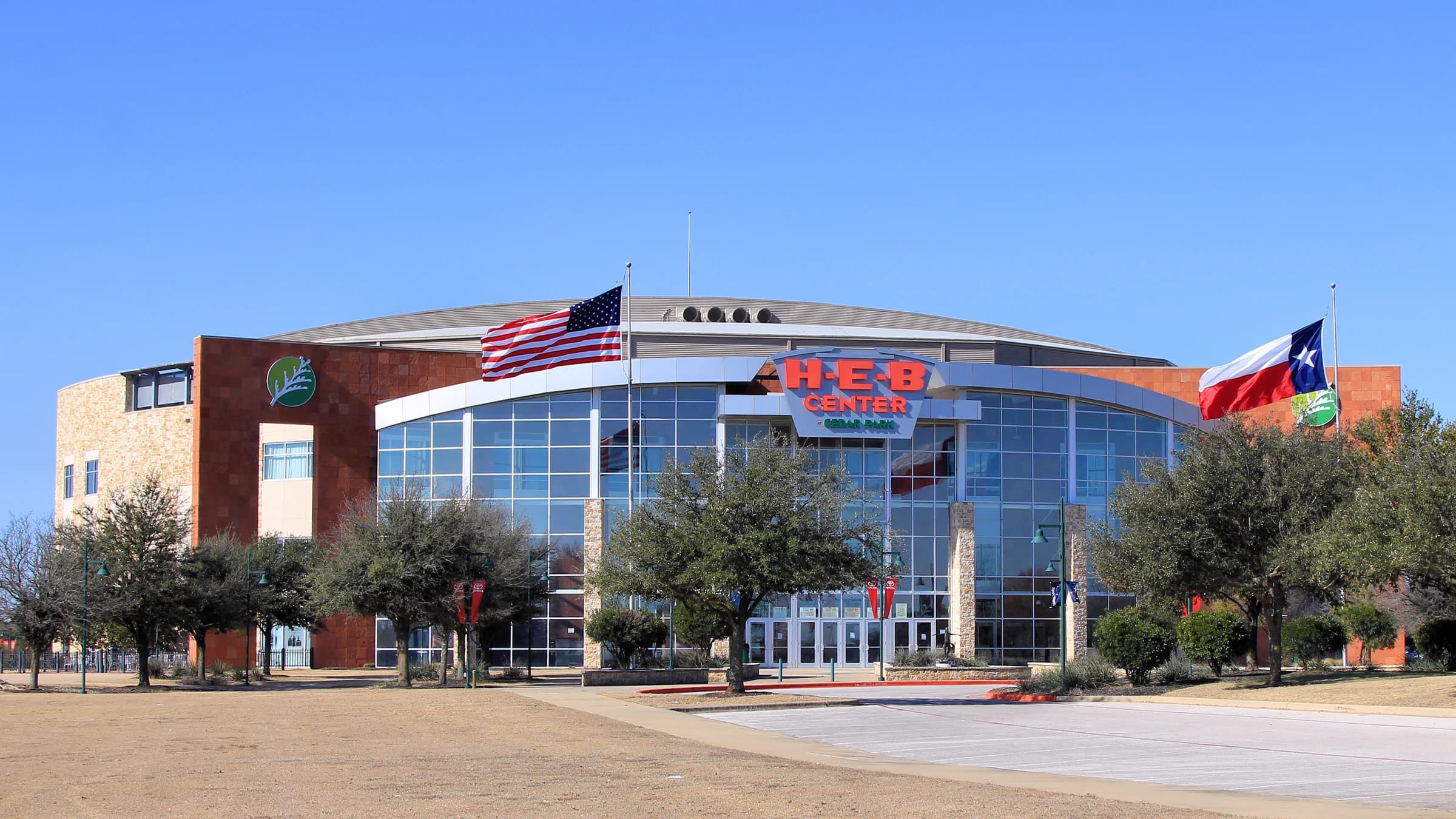 The glass front of the H-E-B Center arena in Cedar Park, Texas, with US and Texas flags and live oaks under a clear blue sky