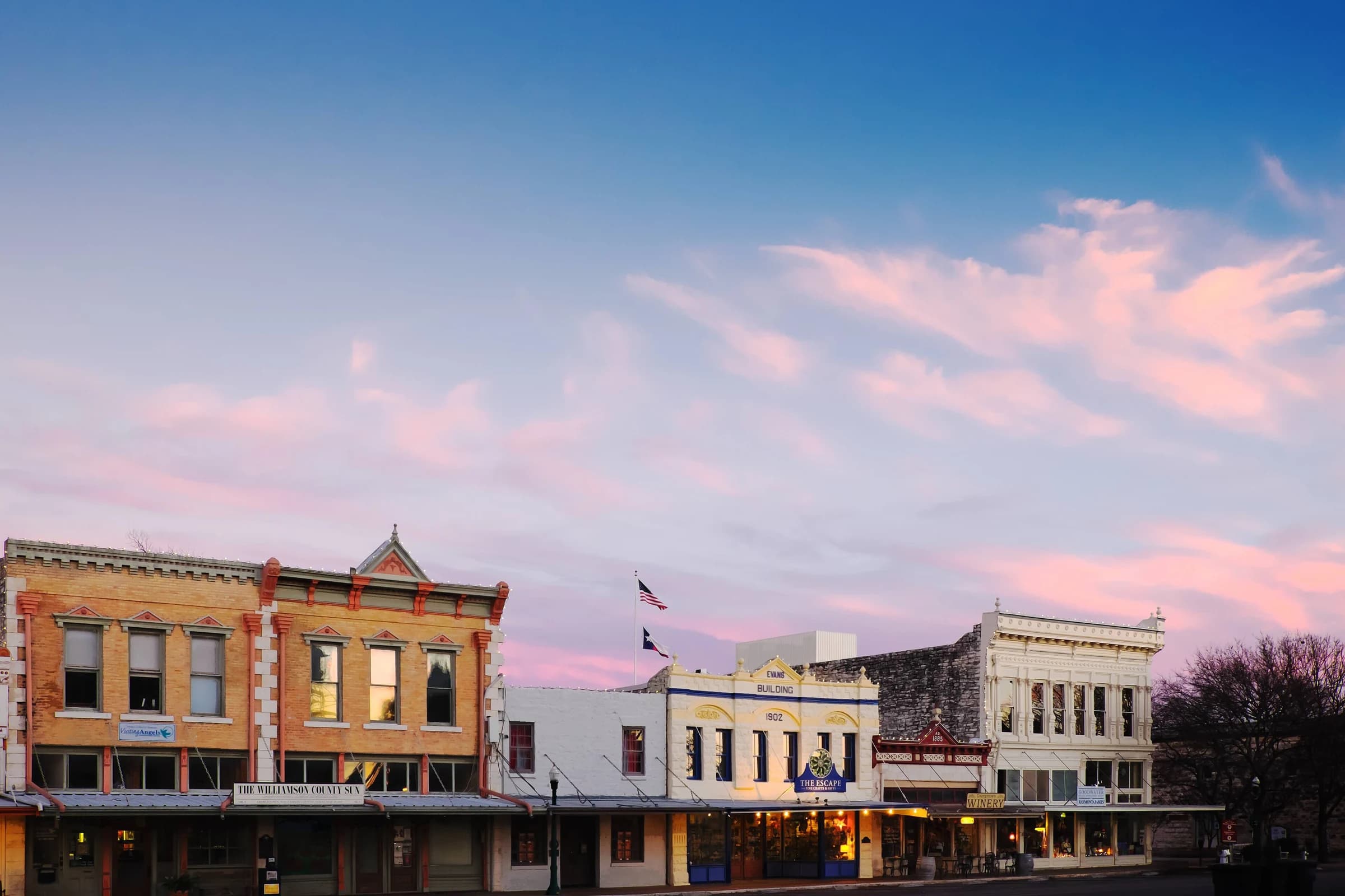 Victorian storefronts along South Main Street on the courthouse square in Georgetown, Texas, under a pink dusk sky with the US and Texas flags flying