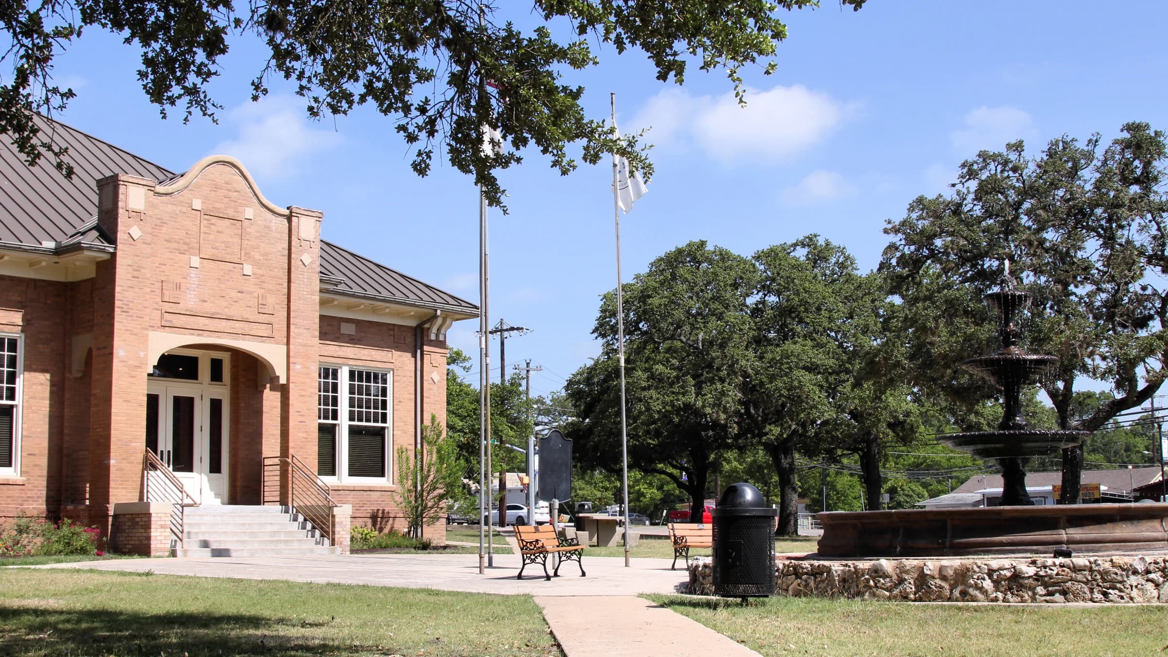 City Square Park in downtown Kyle, Texas, with a tiered fountain, benches and live oaks beside an old red-brick civic building