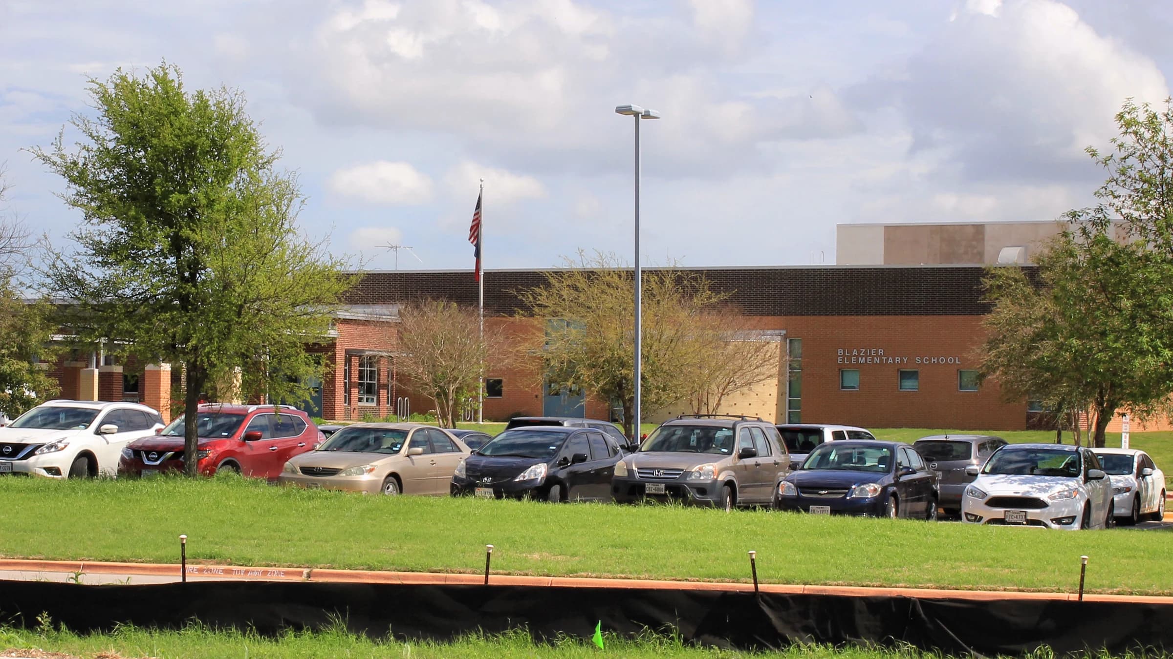 The red-brick front of Blazier Elementary School on Vertex Boulevard in Goodnight Ranch, southeast Austin, with a flagpole, cars in the drop-off lane and a green field in front