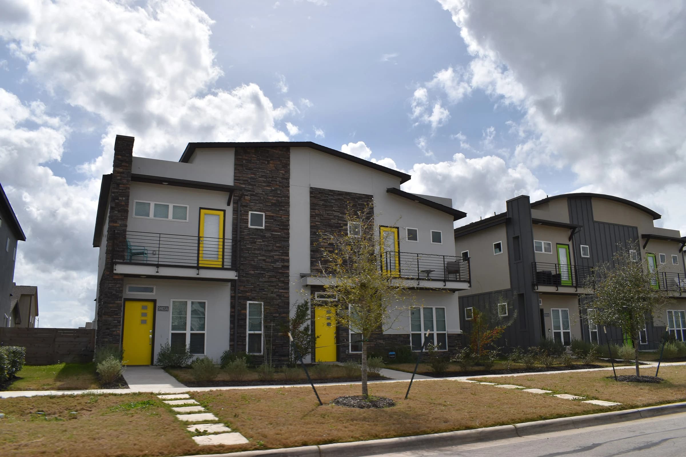 Two-story houses with yellow front doors, stacked-stone chimneys and metal balcony rails along a new street in Goodnight Ranch, southeast Austin, with young trees staked in the front yards
