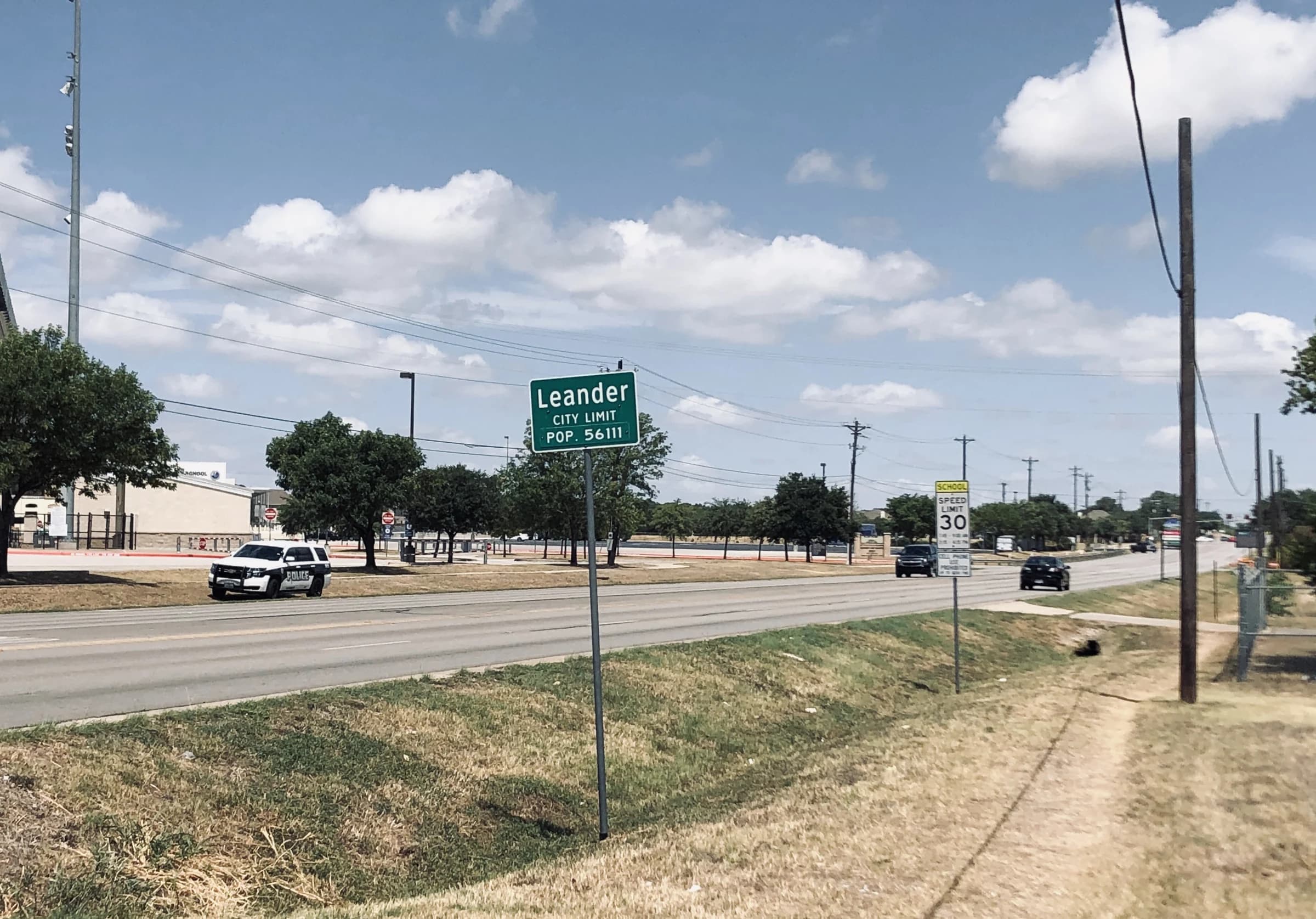 A green Leander city limit sign beside a four-lane road on a sunny day in Leander, Texas, with young trees, utility poles and a school zone speed sign behind it