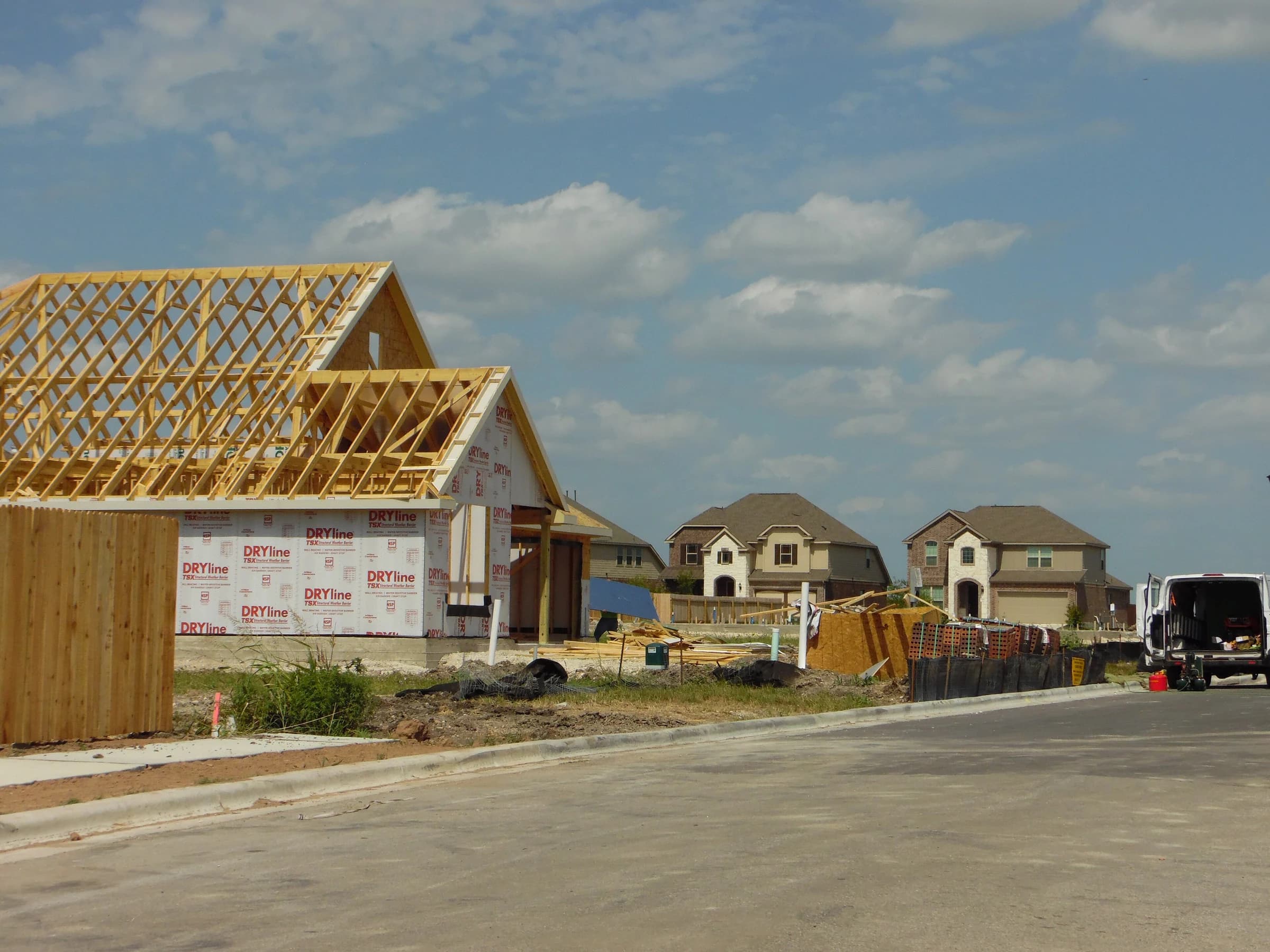 A new house in Pflugerville's Blackhawk subdivision with its roof trusses up and walls wrapped in weather barrier, beside a freshly poured street and finished two-story brick and stone homes