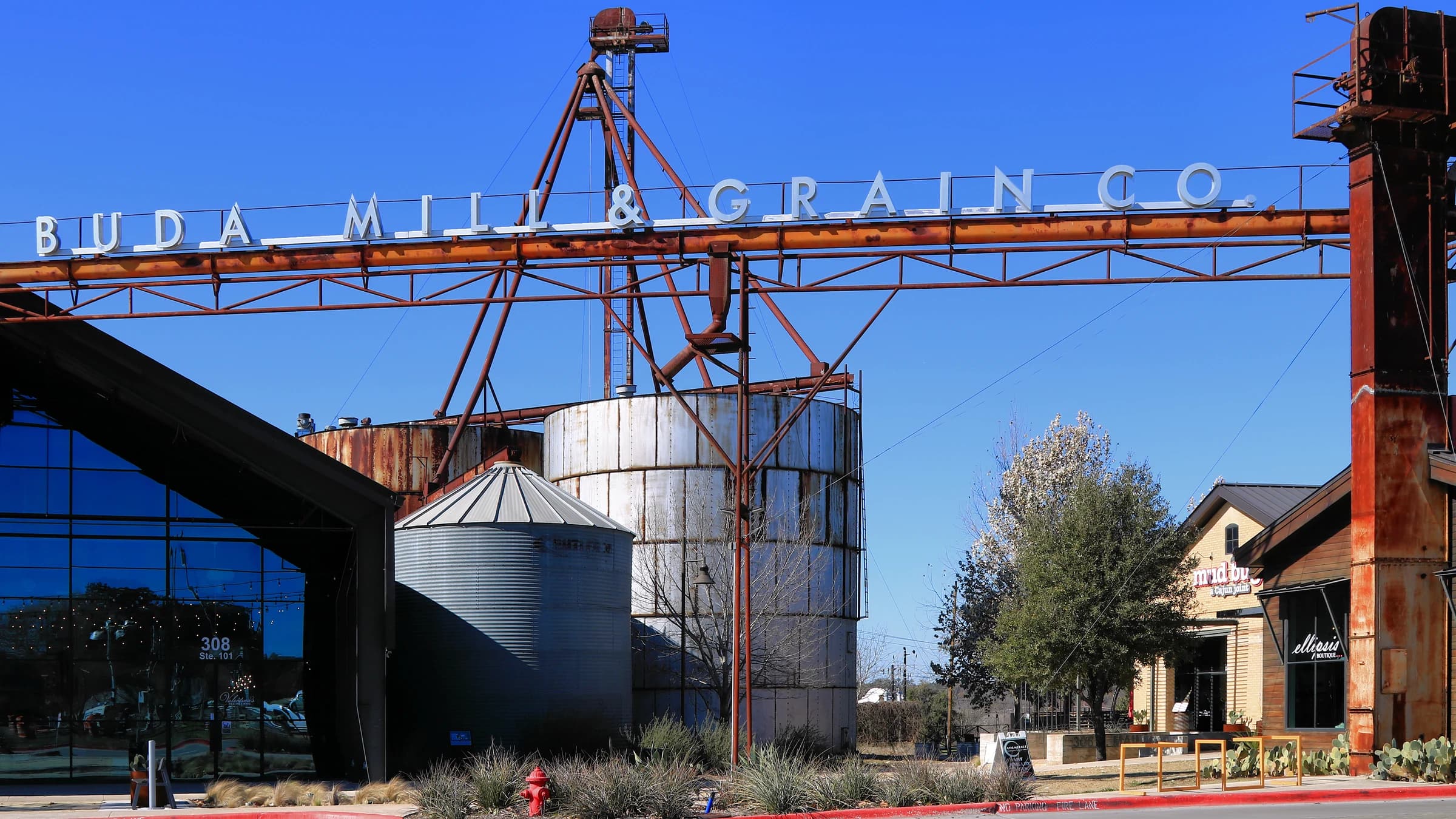 The Buda Mill & Grain Co. shopping center in Buda, Texas, with a rusted steel gantry spelling out its name above old grain silos and a row of shops