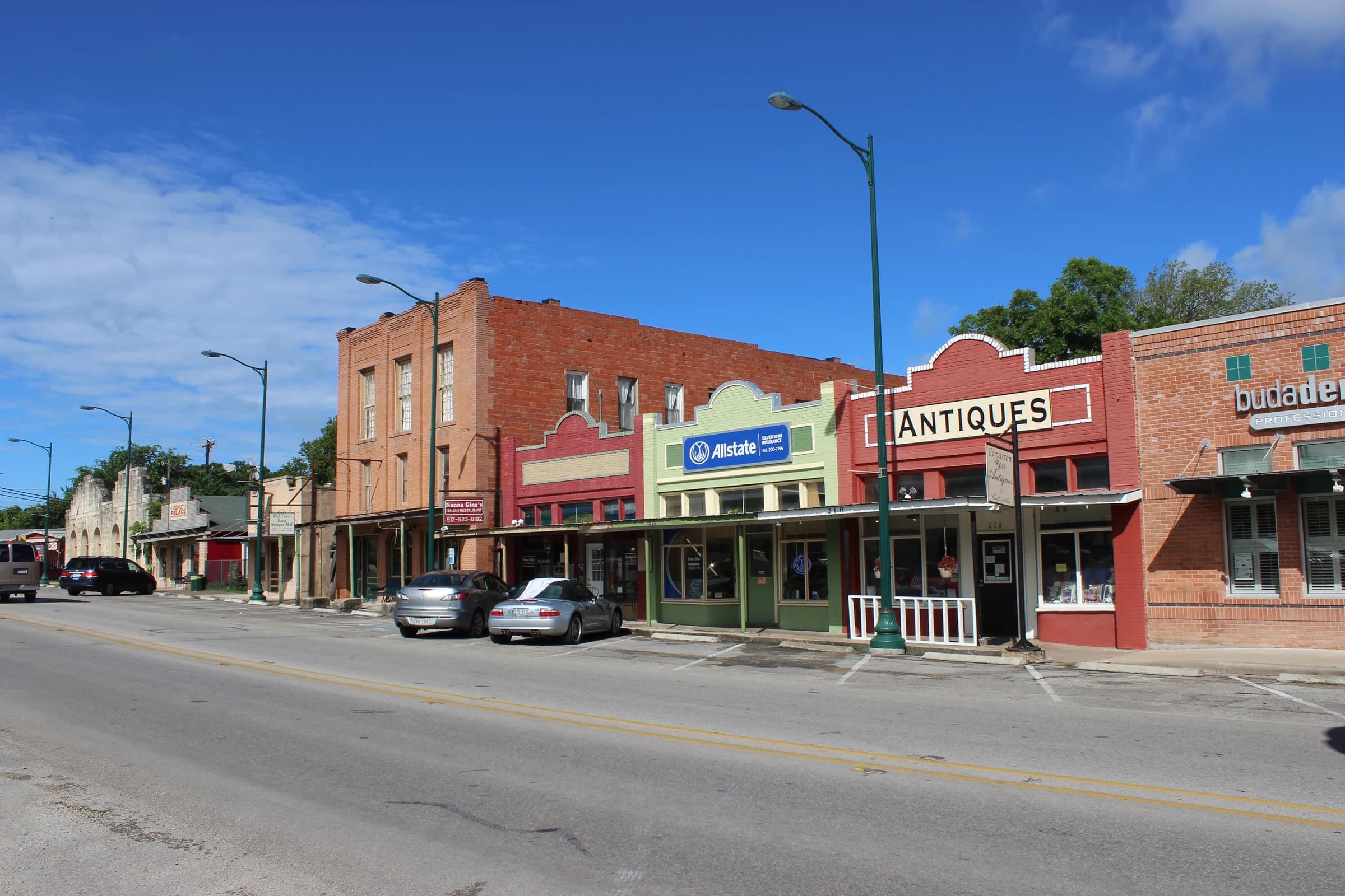 A row of historic storefronts on Main Street in downtown Buda, Texas, with a two-story brick building, green street lamps and parked cars under a blue sky