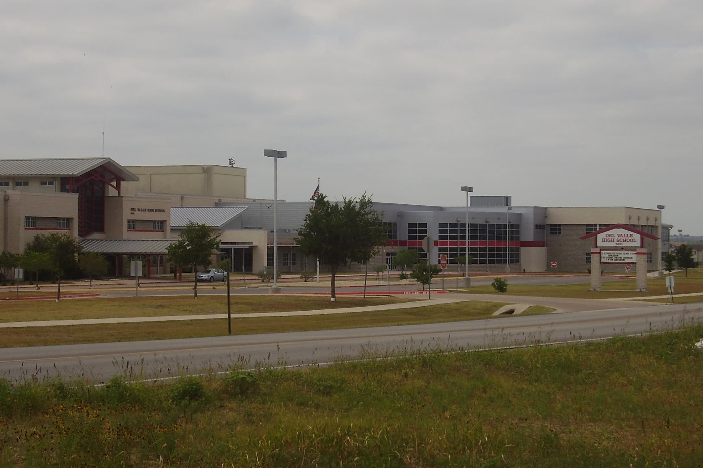 The tan-brick and red-trimmed front of Del Valle High School on Ross Road in Del Valle, Texas, with its campus sign, a flagpole and young trees beyond a grassy roadside