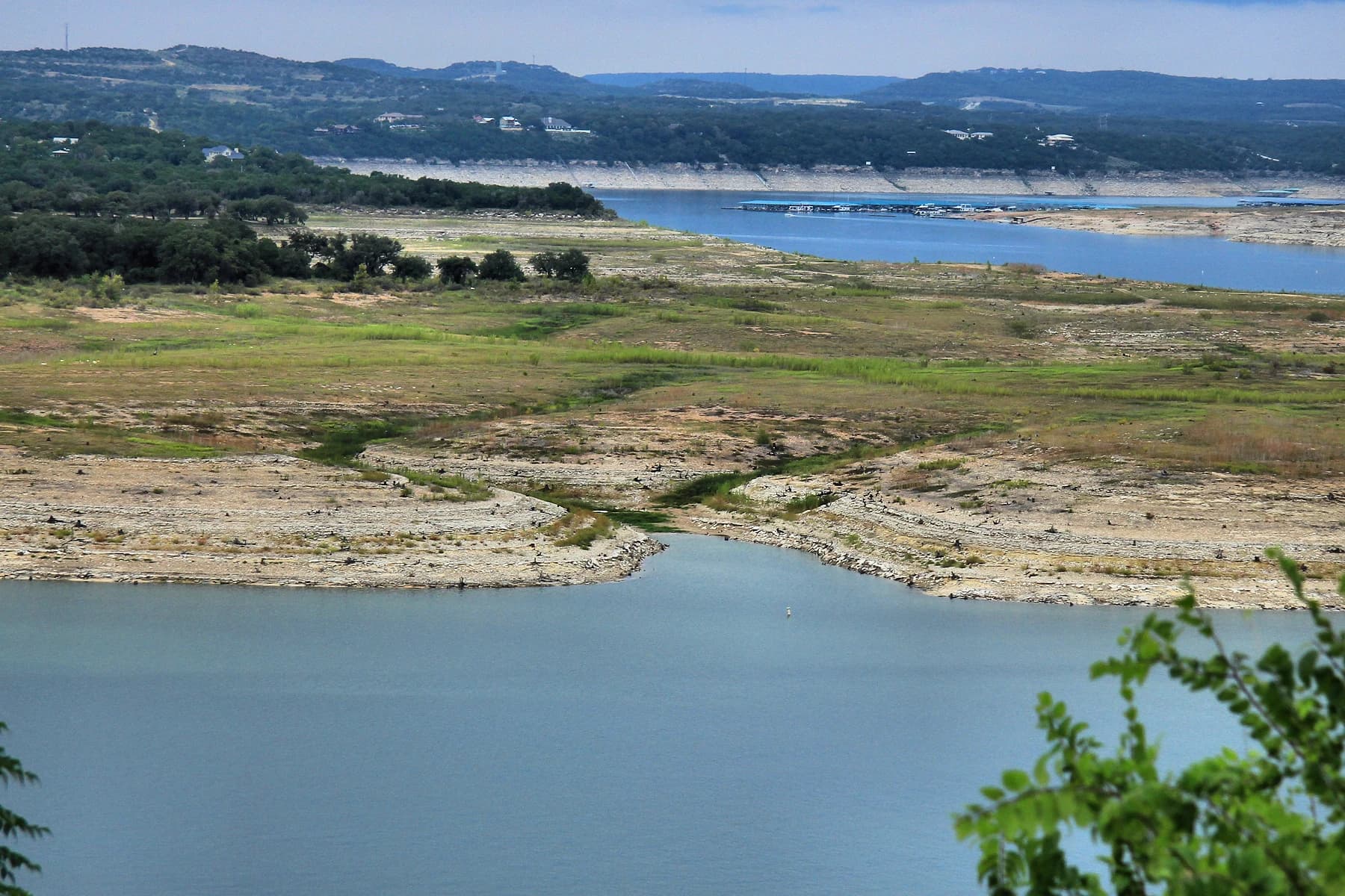 Lake Travis far below its full level during the 2013 drought, with a wide band of bare limestone shoreline and dry cove bottom between the water and the tree line, and a marina floating in the distance
