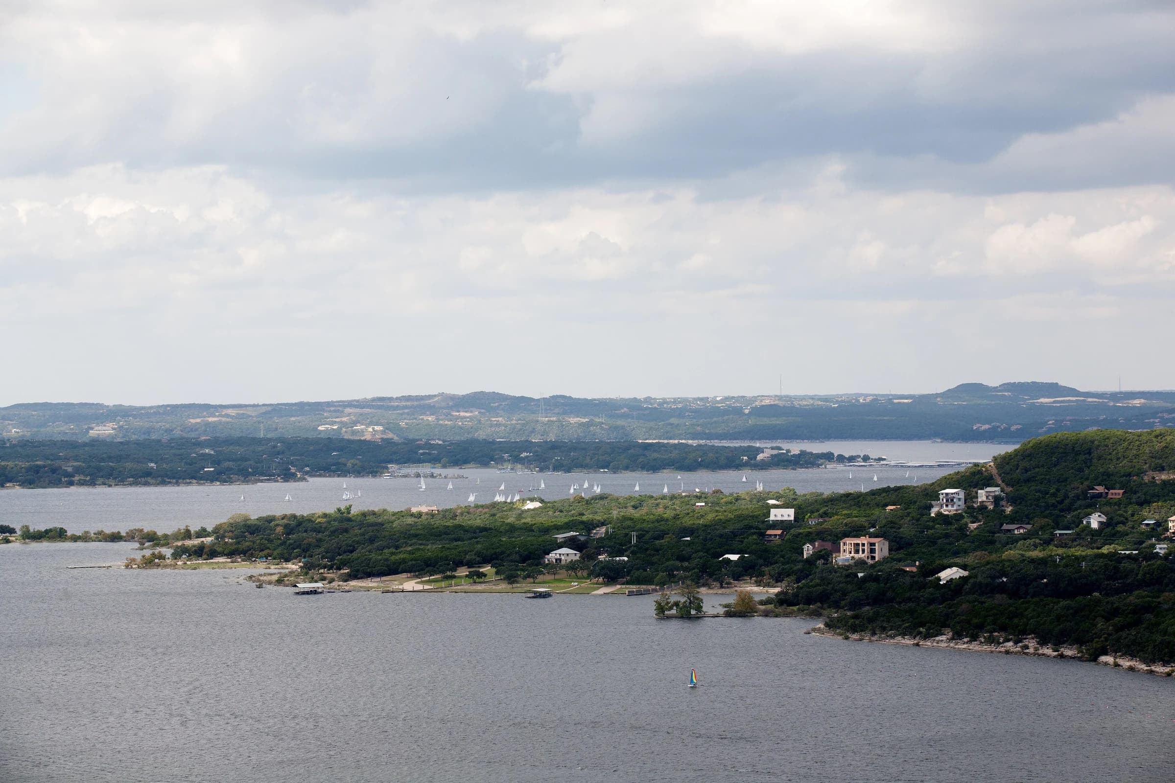 Lake Travis near full pool west of Austin, with sailboats on the water, a wooded point lined with lakeside homes and Hill Country ridges under a cloudy sky