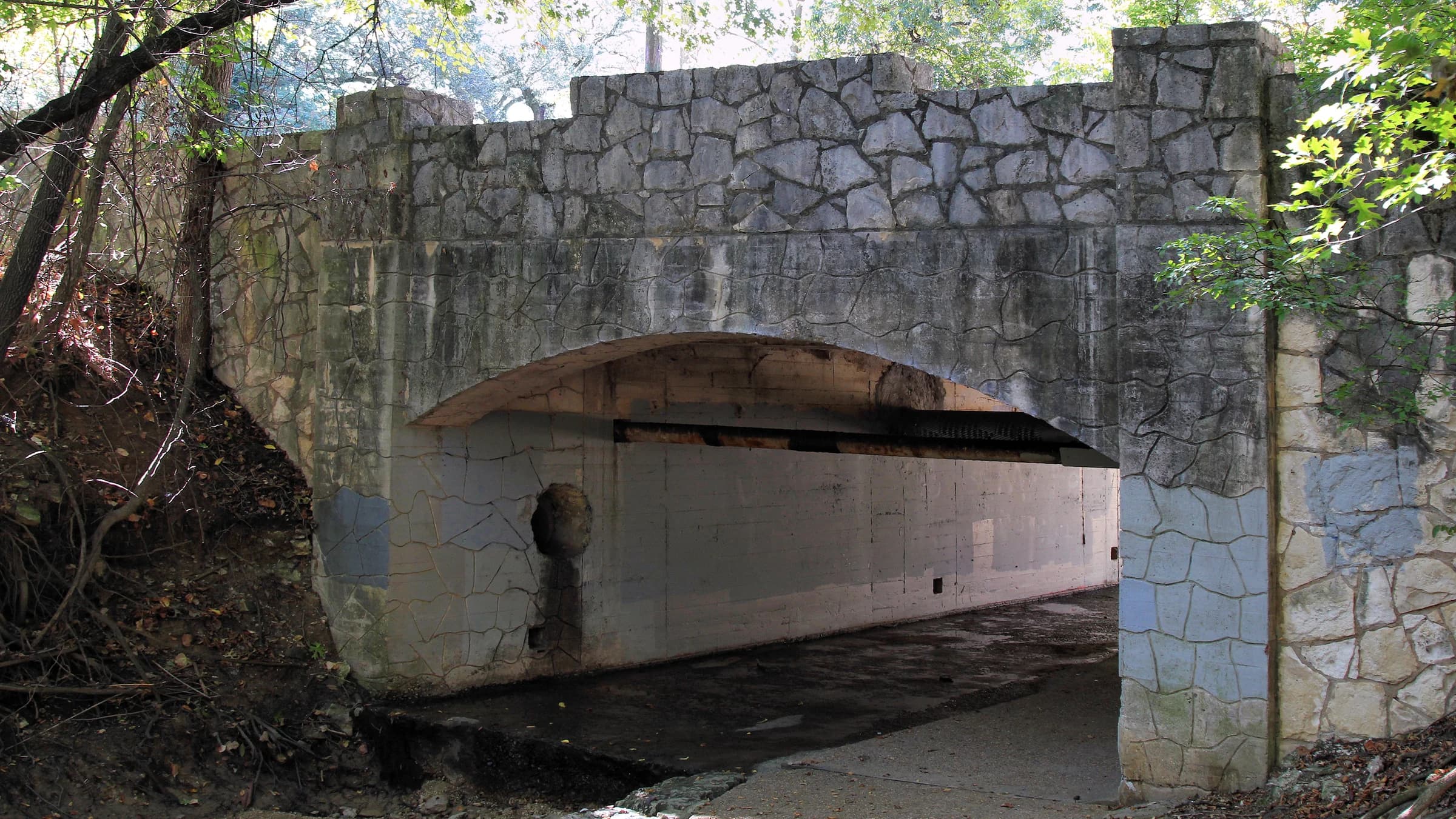 Rustic stone-faced vehicle bridge arching over a shaded path in the Travis Heights-Fairview Park Historic District in South Austin
