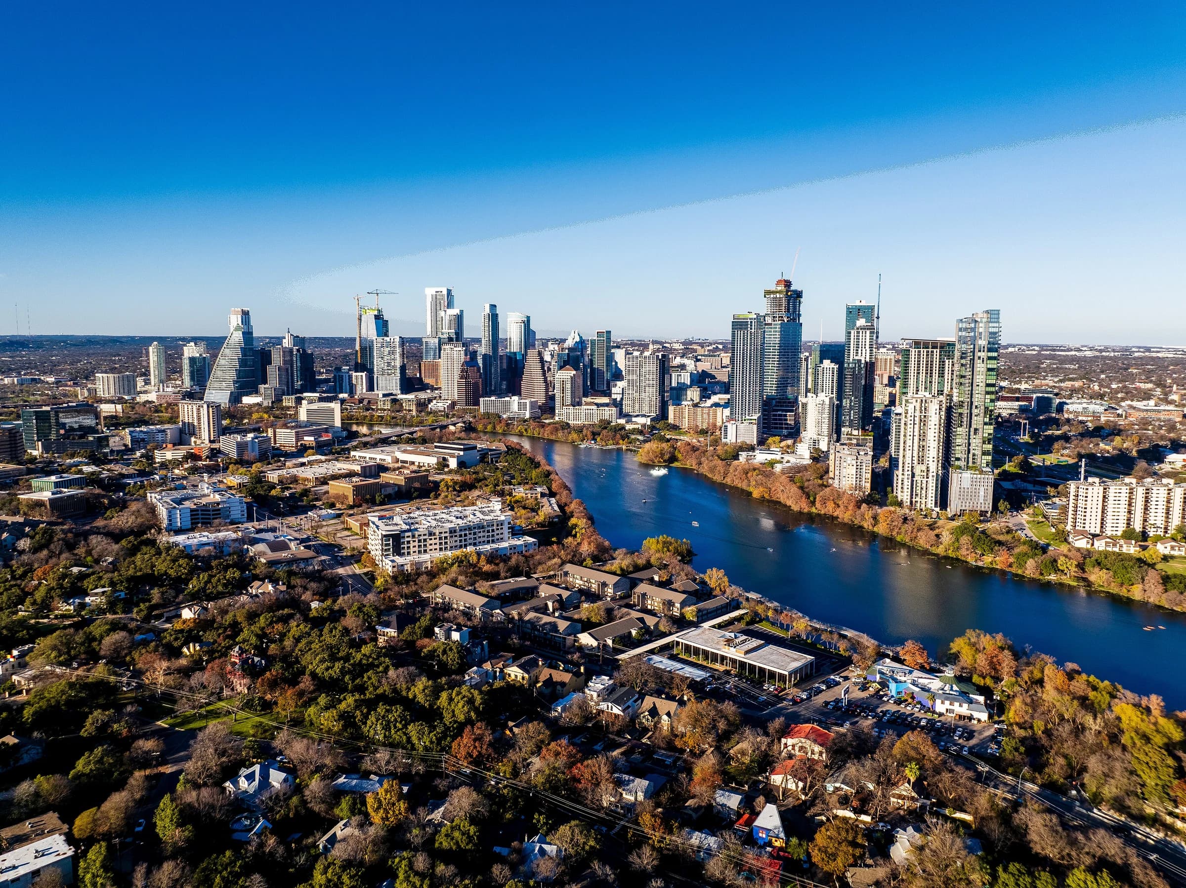 Aerial view north over Travis Heights in South Austin, its houses under autumn trees, with Lady Bird Lake and the downtown towers beyond